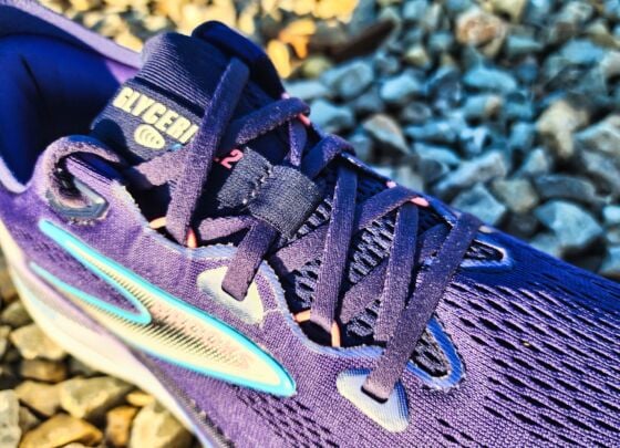 A pair of Brooks Glycerin GTS 22 running shoes with white soles and blue accents are placed outdoors on a sunlit concrete surface, with gravel and greenery in the background—a perfect setting for a quick review.