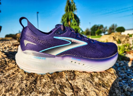A pair of Brooks Glycerin GTS 22 running shoes with white soles and blue accents are placed outdoors on a sunlit concrete surface, with gravel and greenery in the background—a perfect setting for a quick review.