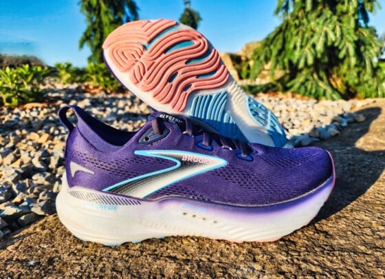 A pair of Brooks Glycerin GTS 22 running shoes with white soles and blue accents are placed outdoors on a sunlit concrete surface, with gravel and greenery in the background—a perfect setting for a quick review.