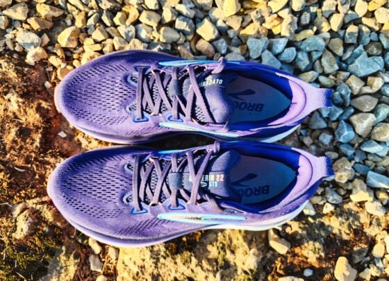 A pair of Brooks Glycerin GTS 22 running shoes with white soles and blue accents are placed outdoors on a sunlit concrete surface, with gravel and greenery in the background—a perfect setting for a quick review.