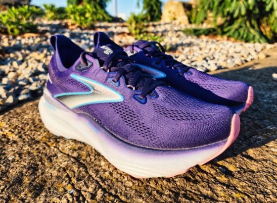 A pair of Brooks Glycerin GTS 22 running shoes with white soles and blue accents are placed outdoors on a sunlit concrete surface, with gravel and greenery in the background—a perfect setting for a quick review.