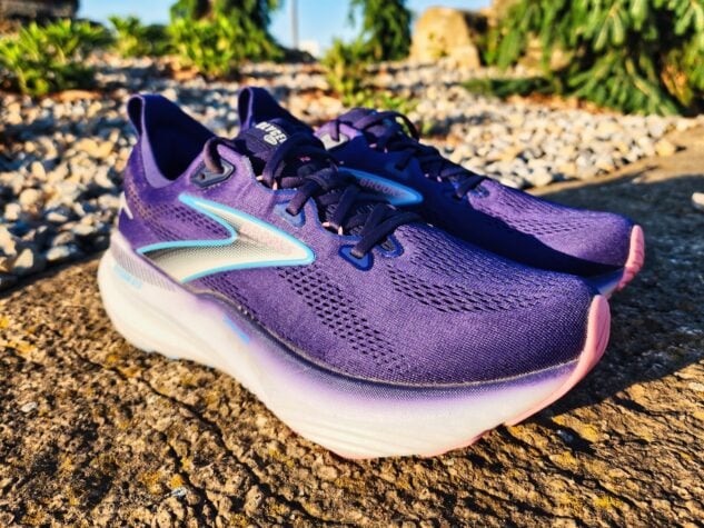 A pair of Brooks Glycerin GTS 22 running shoes with white soles and blue accents are placed outdoors on a sunlit concrete surface, with gravel and greenery in the background—a perfect setting for a quick review.